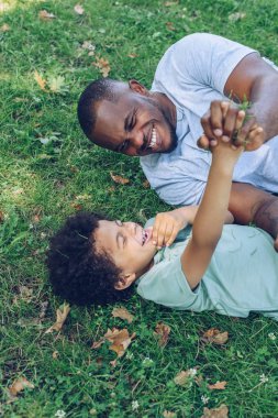 Neşeli Afrikalı Amerikalı baba ve oğul parkta çimenlikte yatarken eğleniyorlar.