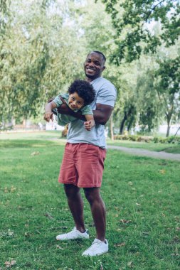 happy african american man holding adorbale son while having fun in park