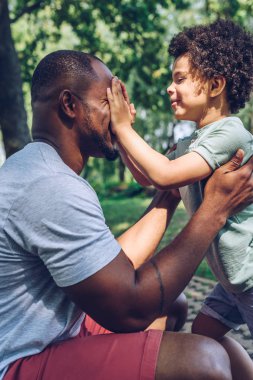 Şirin Afrikalı Amerikalı çocuk parkta eğlenirken babasının gözlerini elleriyle kapatıyor.