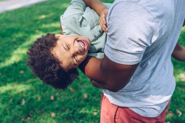 cropped view of african american man holding laughing son while having son in park clipart
