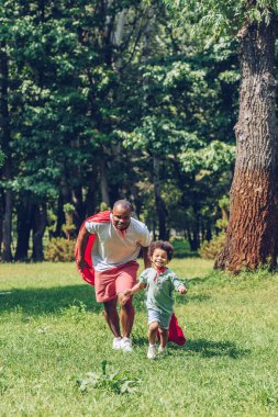 cheerful african american father and son running in costumes of superheroes in park