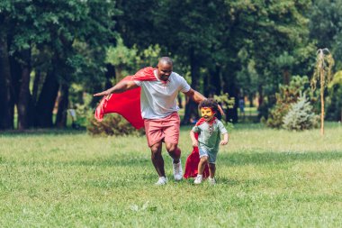 happy african american father and son running in costumes of superheroes in park