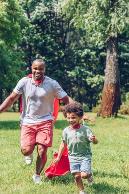 cheerful african american father and son running in costumes of superheroes in park