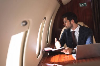 Genç işadamı iş gezisi sırasında uçakta dizüstü bilgisayar ve defterle çalışıyor. 