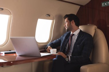 Düşünceli iş adamı iş gezisi sırasında laptopuyla uçakta kahve tutuyor. 