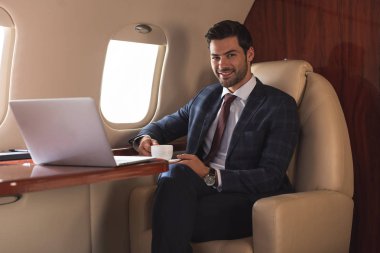 Gülümseyen iş adamı iş gezisi sırasında elinde laptopuyla uçakta bir fincan kahve tutuyor. 