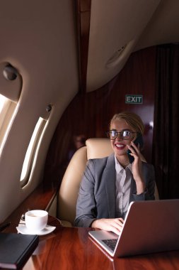 İş gezisi sırasında uçakta akıllı telefon ve dizüstü bilgisayarla çalışan gülümseyen iş kadını 