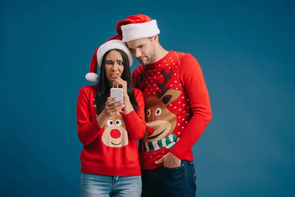 Šokovaný Pár Santa Kloboučky Vánoční Svetry Používající Smartphone Izolovaný Modrém — Stock fotografie