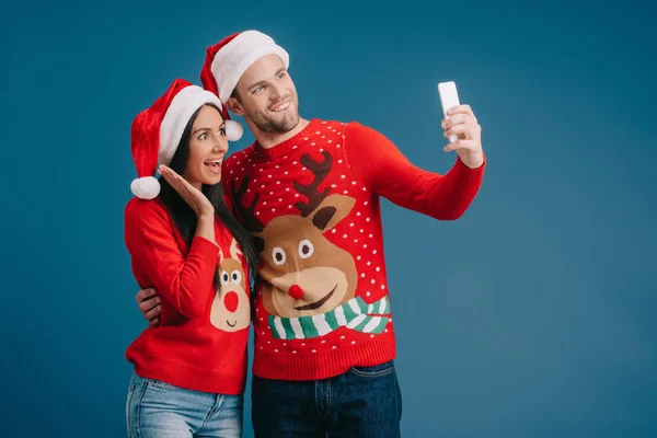 サンタの帽子とクリスマスセーターで興奮したカップルは 青で孤立したスマートフォンで自分撮りを取ります — ストック写真