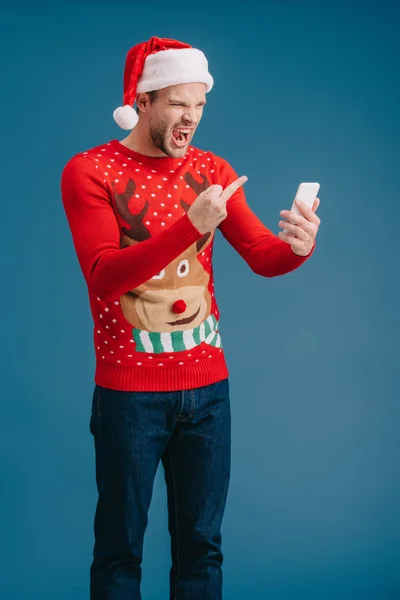 愤怒的男子在圣诞老人的帽子大喊大叫 并显示中指智能手机 隔离在蓝色 — 图库照片