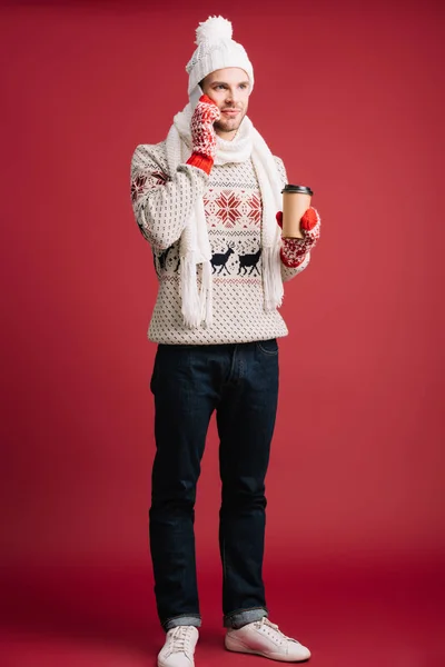 英俊的男子在冬天毛衣 帽子和手套拿着咖啡去 而在智能手机上说话 隔离在红色 — 图库照片