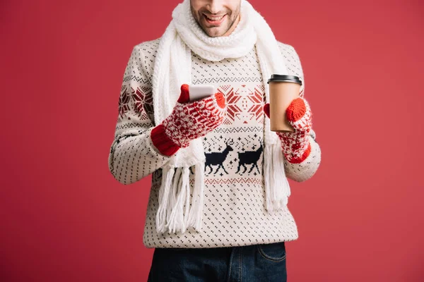 cropped view of man in winter sweater, scarf, hat and mittens holding coffee to go while using smartphone, isolated on red