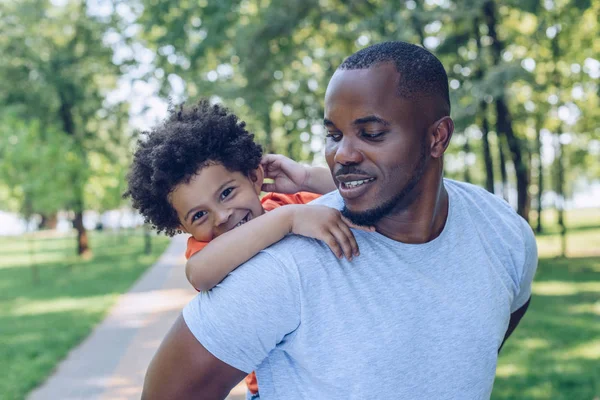 Yakışıklı Afro-Amerikan erkeği parkta neşeli oğlunu sırtında taşıyor.