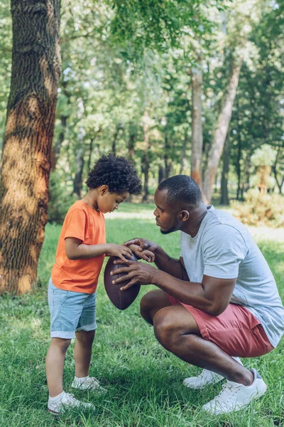 公園で可愛い息子にラグビーボールを見せる若いアフリカ系アメリカ人の父親 — ストック写真