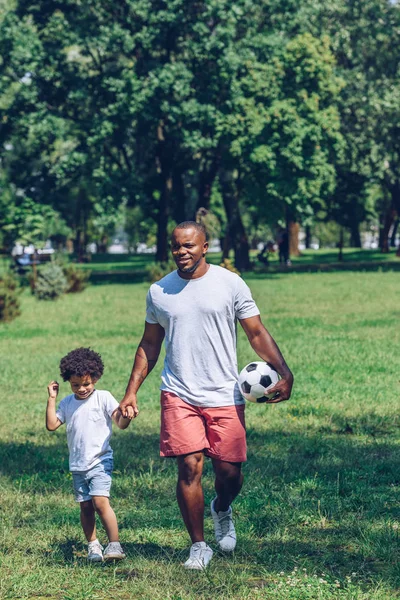Yakışıklı Afro-Amerikalı futbol topuyla parkta yürürken tapılası oğlunun elini tutuyor.