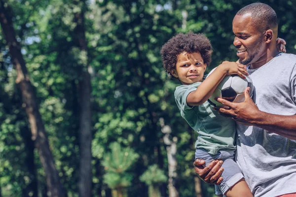 かわいいアフリカ系アメリカ人の少年が父親の手に座ってサッカーボールを持ち — ストック写真