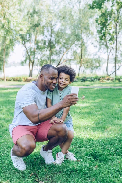 Mutlu Afro-Amerikan baba ve oğlu parkta akıllı telefondan selfie çekiyor.