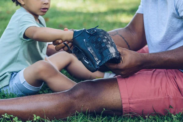 Обрезанный Вид Африканского Американца Показывающего Бейсбольную Перчатку Сыну Сидящего Газоне — стоковое фото