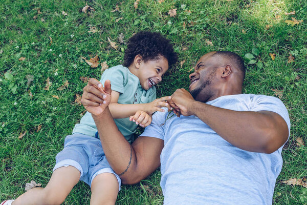happy african american father and son having fun while lying on lawn in park