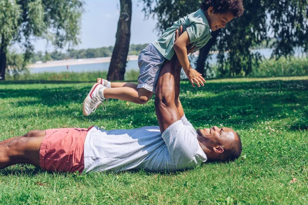 Joven Afroamericano Padre Holding Lindo Hijo Sobre Cabeza Mientras Acostado —  Fotos de Stock