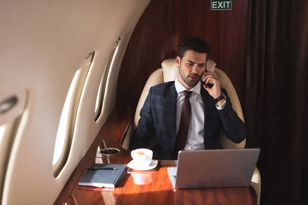 商务旅行期间 男性领导人在使用笔记本电脑的飞机上用智能手机交谈 — 图库照片