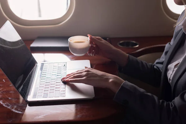 Ausgeschnittene Ansicht einer Geschäftsfrau, die eine Kaffeetasse hält und während einer Geschäftsreise am Laptop im Flugzeug arbeitet — Stockbild Ausgeschnittene Ansicht Einer Geschäftsfrau Die Eine Kaffeetasse Hält Und Während — Stockfoto