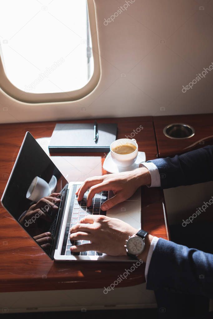 cropped view of businessman working with laptop in plane during business trip 