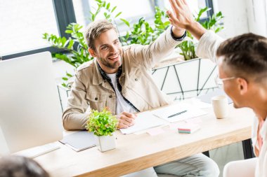 young, cheerful businessmen giving high five while sitting at workplace in office clipart