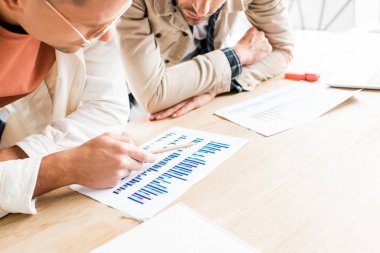 cropped view of two young businessmen analyzing documents with graphs and charts while working on startup project together in office clipart