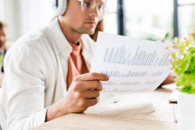 selective focus of young businessman sitting in headphones and analyzing paper with graphs and charts clipart