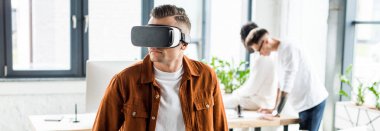 Çok kültürlü meslektaşlar ofiste çalışırken genç iş adamının VR kulaklığı kullanışının panoramik görüntüsü