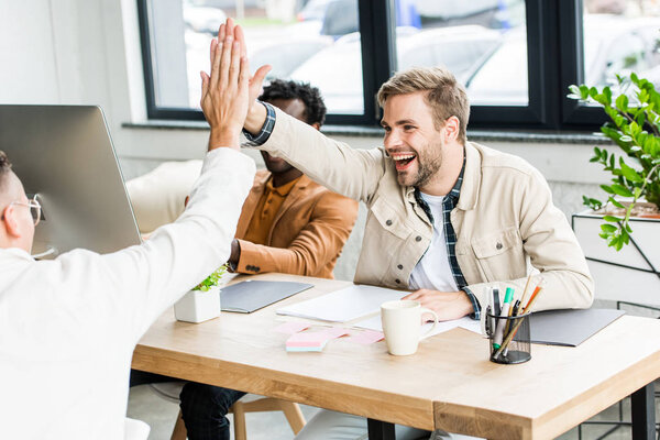 young, cheerful businessmen giving high five while sitting at workplace in office