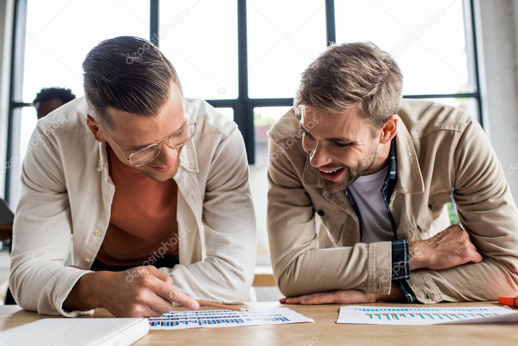 two young smiling businessmen analyzing papers with graphs and charts while working on startup project together in office 