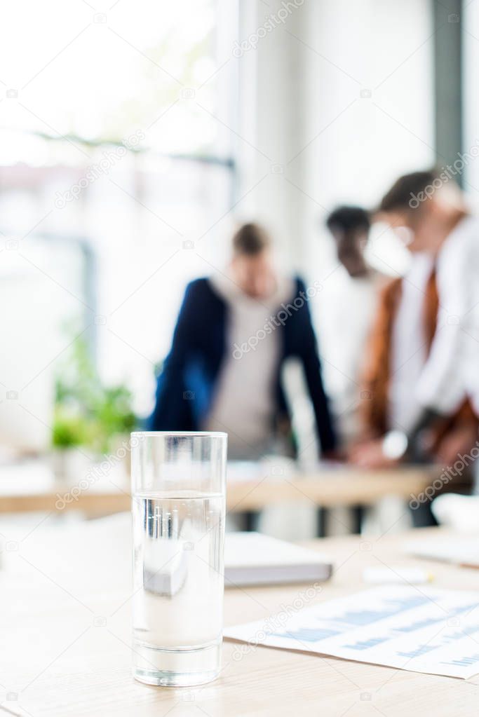 selective focus of glass with water on desk near businesspeople standing at workplace in office