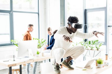 Afro-Amerikan iş adamı VR kulaklık kullanırken masaya çömeliyor ve iş arkadaşları ofiste çalışıyor.