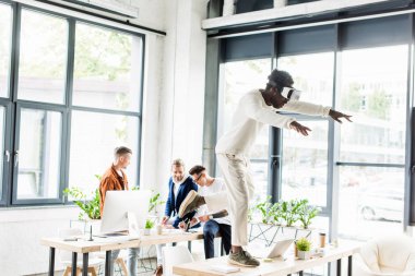 Afrika kökenli Amerikalı iş adamı VR cihazını kullanıyor ve meslektaşları ofiste çalışırken masasında duruyor.