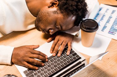 Yorgun Afrikalı Amerikalı iş adamı laptopun yanında uyuyor ve geceleri ofisinde tek kullanımlık bardakta.