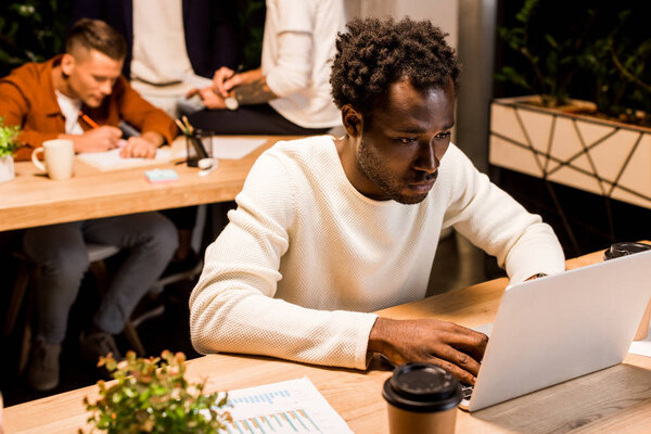 exhausted african american businessman using laptop while working at night in office near colleagues