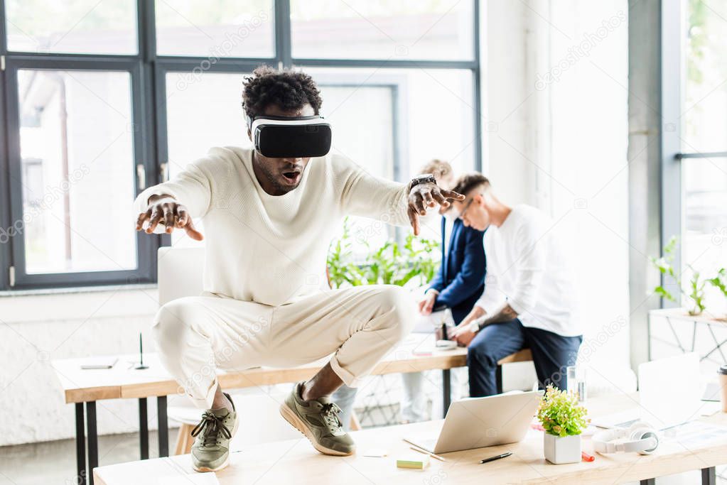 african american businessman using vr headset and squatting on desk while colleagues working in office