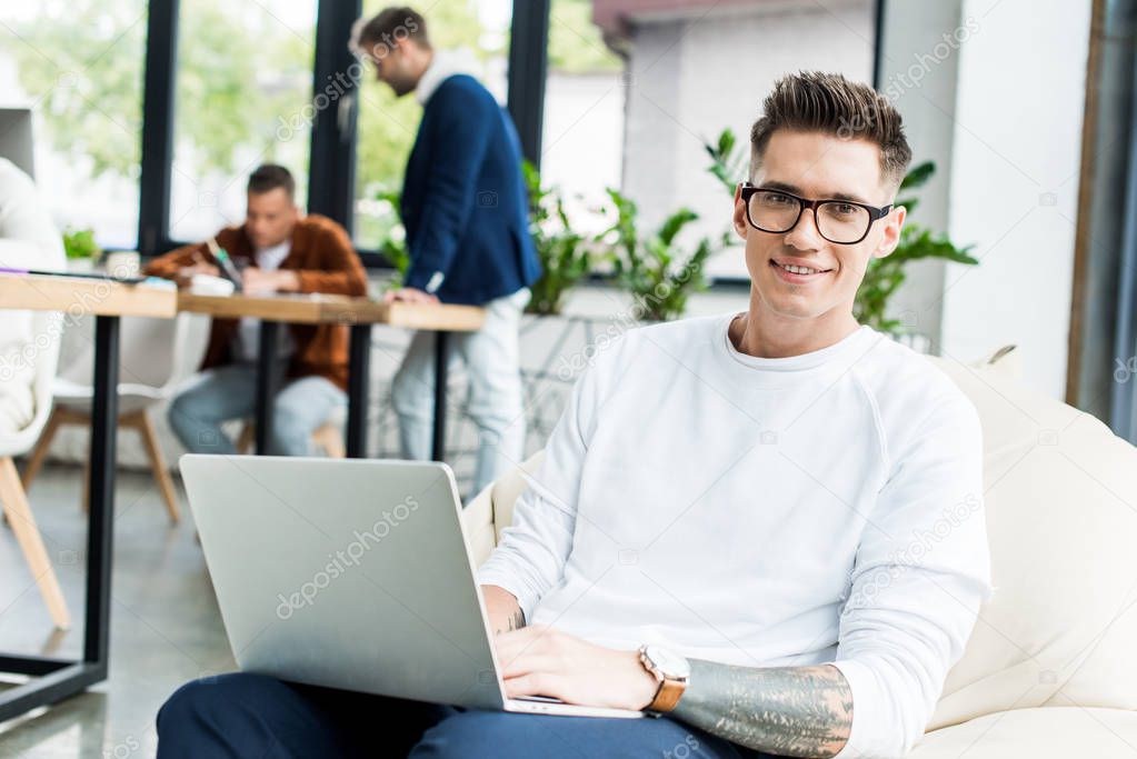 young businessman using laptop and smiling at camera while working near colleagues in office