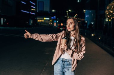 pembe ceket otostop çekici kadın ve gece şehirde kağıt bardak tutarak 