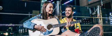 panoramic shot of handsome boyfriend with plastic cup and attractive girlfriend playing acoustic guitar in night city  clipart