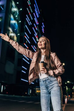 pembe ceket otostop çekici kadın ve gece şehirde kağıt bardak tutarak  