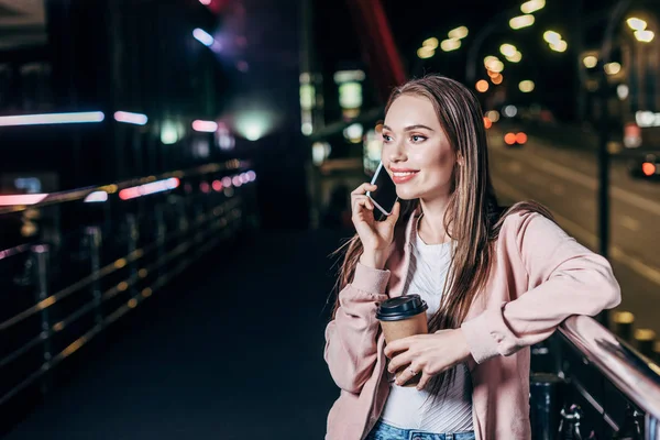 有吸引力的女人在粉红色的夹克说话的智能手机和持有纸杯在夜城 — 图库照片