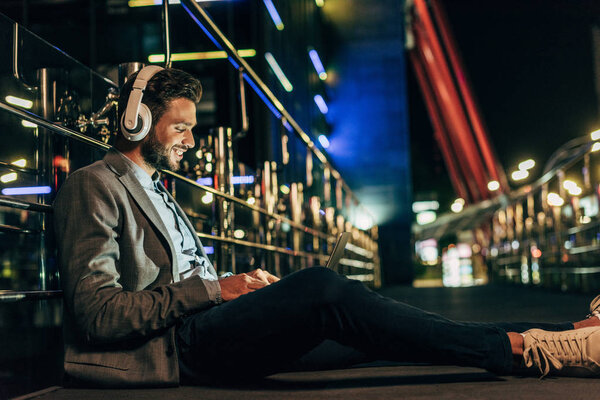 красивый бизнесмен в формальной одежде с наушниками сидя и используя ноутбук в ночном городе  