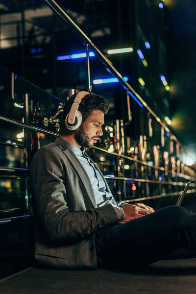красивый бизнесмен в формальной одежде с наушниками сидя и используя ноутбук в ночном городе  
