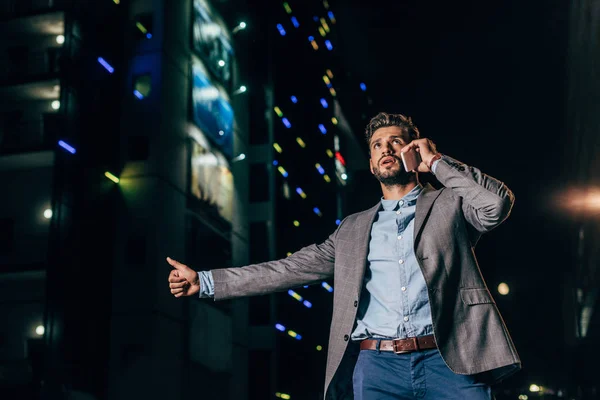 stock image handsome businessman in formal wear talking on smartphone and thumbing in night city 