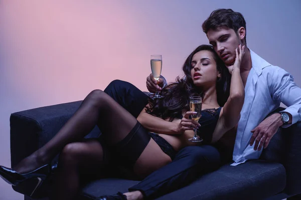 穿着黑色内衣裤的迷人女孩 戴着香槟酒杯躺在紫色背景的沙发上的英俊男人 — 图库照片