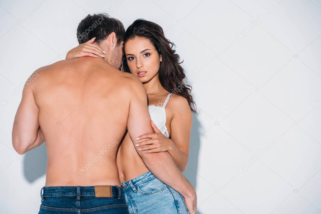 back view of shirtless young man hugging sexy girl in white bra on grey background
