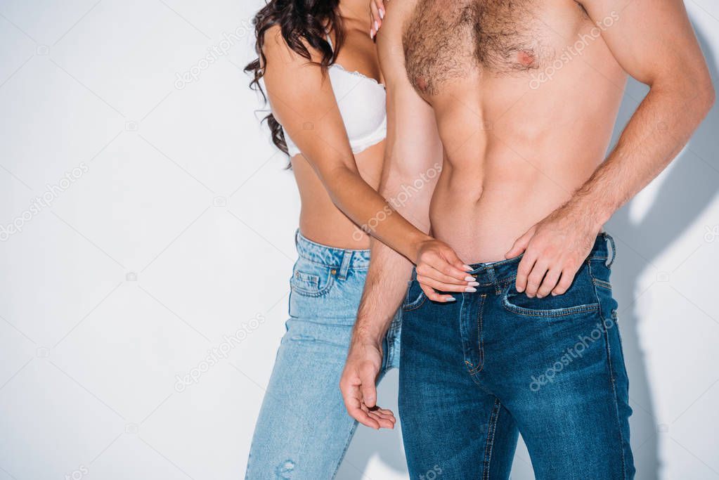 cropped view of girl in white bra unbuttoning jeans of boyfriend on grey background